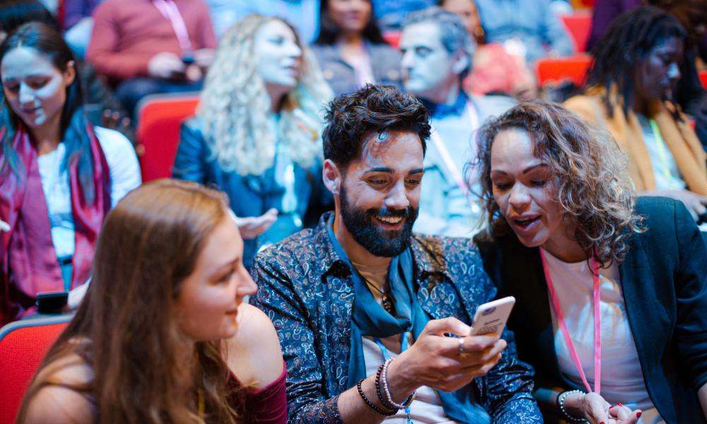 Three attendees at a crowded conference smile while looking at a smartphone as others sit and talk in the background.