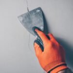 A close-up of a worker's hand wearing orange gloves and applying a sealant with a spatula over a crack in concrete.