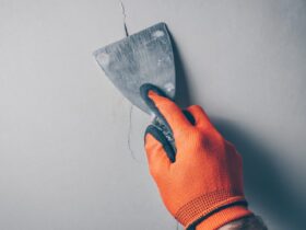 A close-up of a worker's hand wearing orange gloves and applying a sealant with a spatula over a crack in concrete.