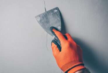 A close-up of a worker's hand wearing orange gloves and applying a sealant with a spatula over a crack in concrete.