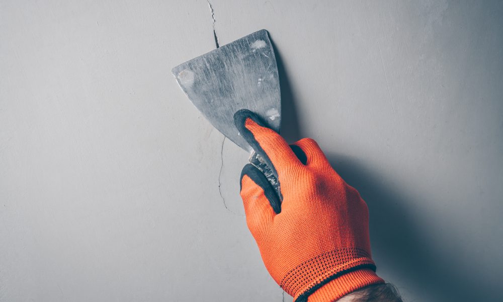 A close-up of a worker's hand wearing orange gloves and applying a sealant with a spatula over a crack in concrete.