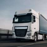 A semi truck with a cargo trailer driving along a highway with motion blur visible on the road and wheels.