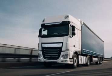 A semi truck with a cargo trailer driving along a highway with motion blur visible on the road and wheels.
