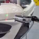 A metrology laser measures a white bumper on a vehicle. There is a green dot and a red verticle line on the bumper.