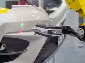 A metrology laser measures a white bumper on a vehicle. There is a green dot and a red verticle line on the bumper.