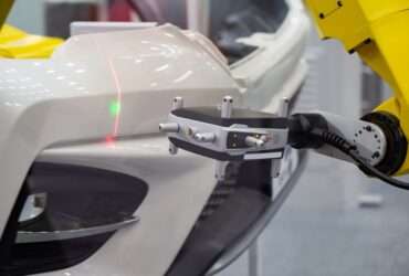 A metrology laser measures a white bumper on a vehicle. There is a green dot and a red verticle line on the bumper.