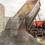 A dump truck unloads a pile of gravel at a construction site, with buildings and cranes in the background.