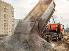 A dump truck unloads a pile of gravel at a construction site, with buildings and cranes in the background.