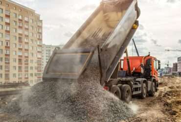A dump truck unloads a pile of gravel at a construction site, with buildings and cranes in the background.