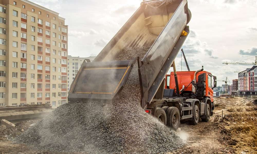 A dump truck unloads a pile of gravel at a construction site, with buildings and cranes in the background.