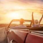A couple driving a classic, convertible car down a dirt road at sunset, with the woman raising her arms in the air.