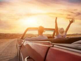 A couple driving a classic, convertible car down a dirt road at sunset, with the woman raising her arms in the air.