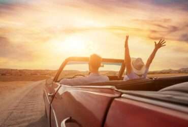 A couple driving a classic, convertible car down a dirt road at sunset, with the woman raising her arms in the air.