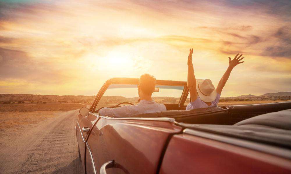 A couple driving a classic, convertible car down a dirt road at sunset, with the woman raising her arms in the air.