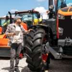 A man with a tablet walks through rows of large, industrial tractors and equipment with large tires and orange accents.
