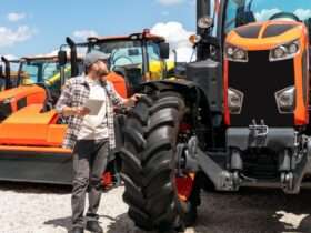 A man with a tablet walks through rows of large, industrial tractors and equipment with large tires and orange accents.