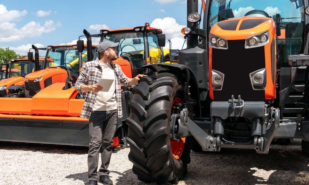A man with a tablet walks through rows of large, industrial tractors and equipment with large tires and orange accents.