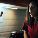 A startled woman in a dark kitchen recoils at a fake spider hanging from the upper cabinets.