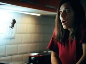 A startled woman in a dark kitchen recoils at a fake spider hanging from the upper cabinets.