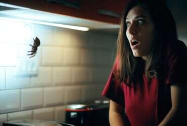 A startled woman in a dark kitchen recoils at a fake spider hanging from the upper cabinets.