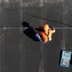 An aerial view of a worker in an orange shirt and hard hat installing a rod on a flat black commercial roof.