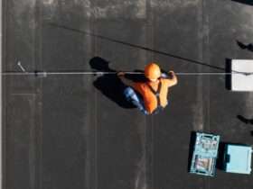 An aerial view of a worker in an orange shirt and hard hat installing a rod on a flat black commercial roof.
