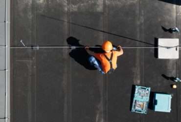 An aerial view of a worker in an orange shirt and hard hat installing a rod on a flat black commercial roof.