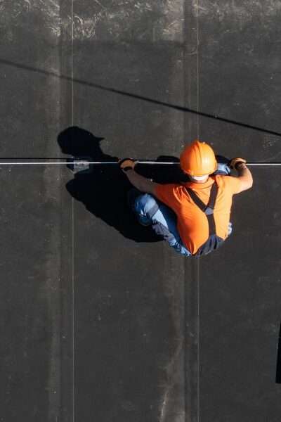 An aerial view of a worker in an orange shirt and hard hat installing a rod on a flat black commercial roof.