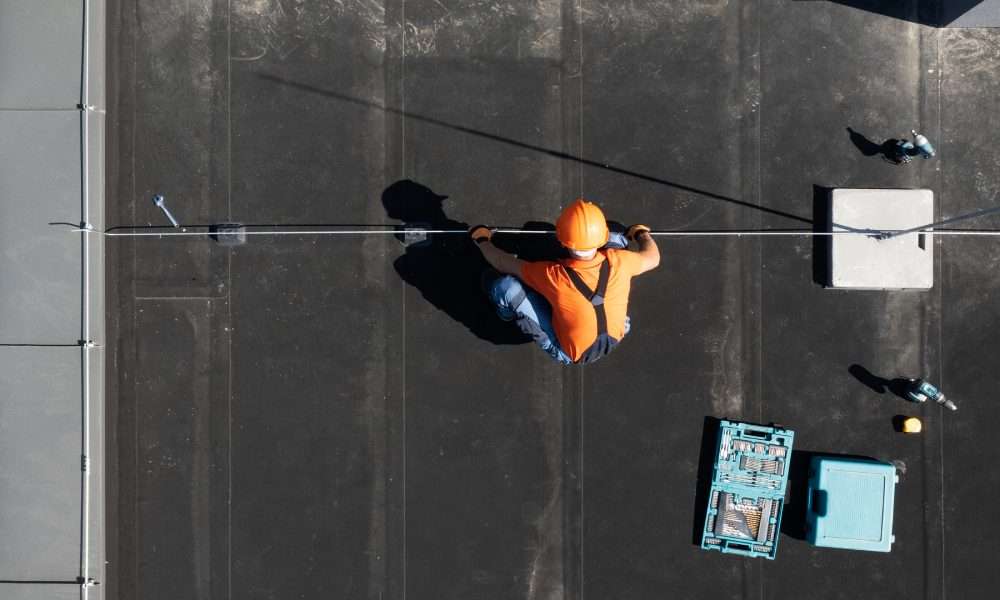 An aerial view of a worker in an orange shirt and hard hat installing a rod on a flat black commercial roof.