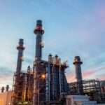 A brightly lit refinery plant with pipes, towers, tanks, and smoke stacks glowing against a dark night sky.