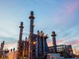 A brightly lit refinery plant with pipes, towers, tanks, and smoke stacks glowing against a dark night sky.