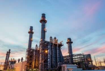 A brightly lit refinery plant with pipes, towers, tanks, and smoke stacks glowing against a dark night sky.