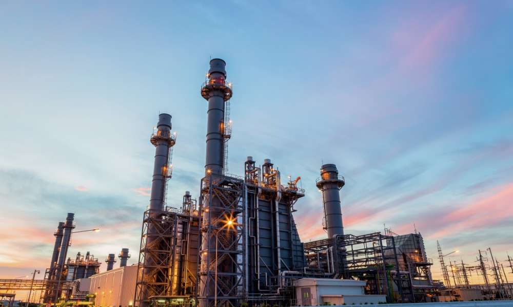 A brightly lit refinery plant with pipes, towers, tanks, and smoke stacks glowing against a dark night sky.
