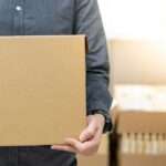 A close-up view shows a person wearing a gray button-down shirt holding a cardboard box. The background is blurry.