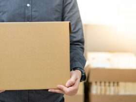 A close-up view shows a person wearing a gray button-down shirt holding a cardboard box. The background is blurry.