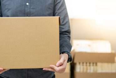 A close-up view shows a person wearing a gray button-down shirt holding a cardboard box. The background is blurry.