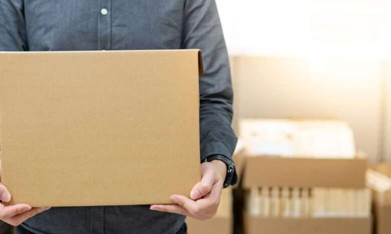 A close-up view shows a person wearing a gray button-down shirt holding a cardboard box. The background is blurry.