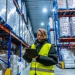 A woman in a cold storage warehouse checking on the inventory with a tablet. She is wearing a heavy coat to keep warm.