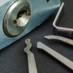 Close-up of a padlock sitting on a table, with its keyhole visible. Three silver metal picks sit next to it.