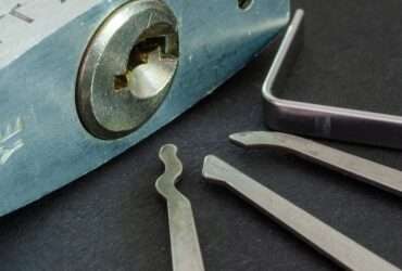 Close-up of a padlock sitting on a table, with its keyhole visible. Three silver metal picks sit next to it.