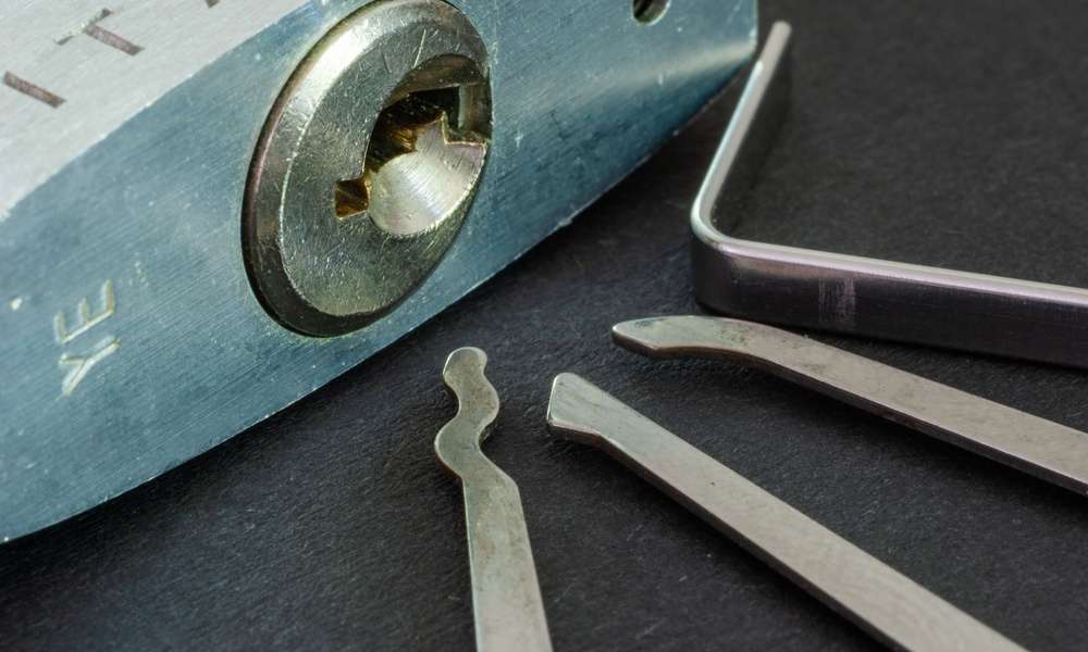 Close-up of a padlock sitting on a table, with its keyhole visible. Three silver metal picks sit next to it.