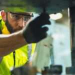 An industrial employee works on a machine. He wears a hard hat, safety glasses, a yellow vest, and a safety glove.