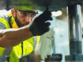 An industrial employee works on a machine. He wears a hard hat, safety glasses, a yellow vest, and a safety glove.