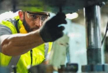An industrial employee works on a machine. He wears a hard hat, safety glasses, a yellow vest, and a safety glove.