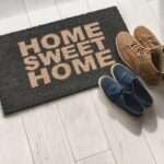 A pair of navy blue sneakers and a pair of brown boots sit on a mat on a tile floor that reads "Home Sweet Home."