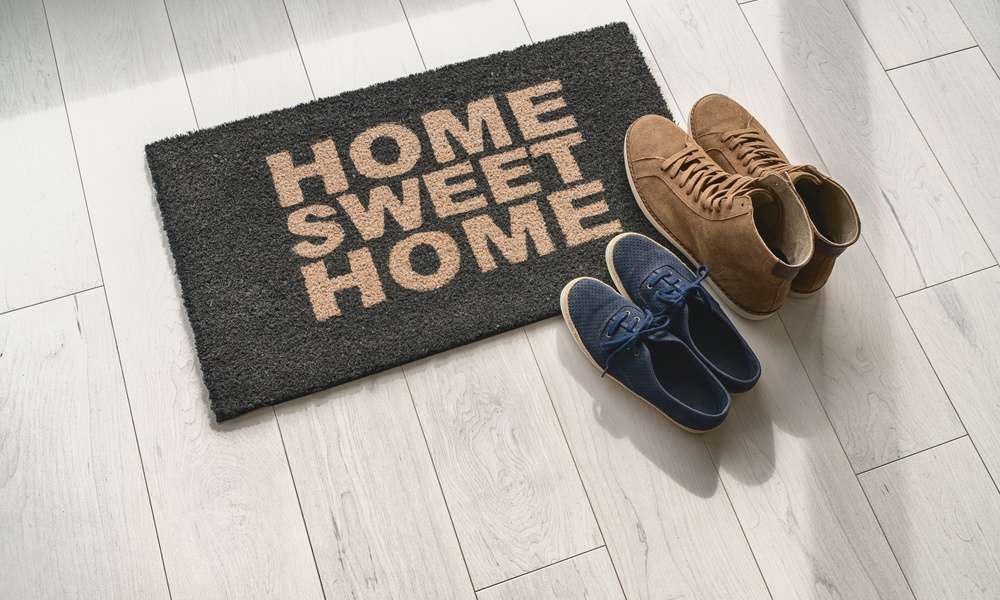 A pair of navy blue sneakers and a pair of brown boots sit on a mat on a tile floor that reads "Home Sweet Home."