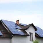 A residential home with multiple solar panel arrays installed across a sloped roof under a clear blue sky.