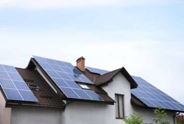 A residential home with multiple solar panel arrays installed across a sloped roof under a clear blue sky.