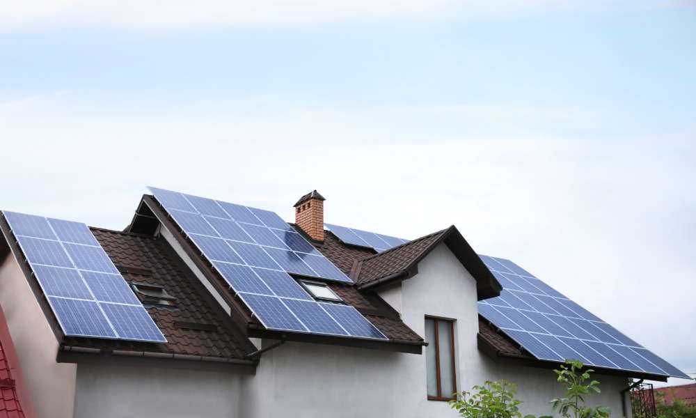A residential home with multiple solar panel arrays installed across a sloped roof under a clear blue sky.