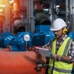 A worker in a hard hat and safety vest checks a clipboard next to a pressure gauge on industrial piping.